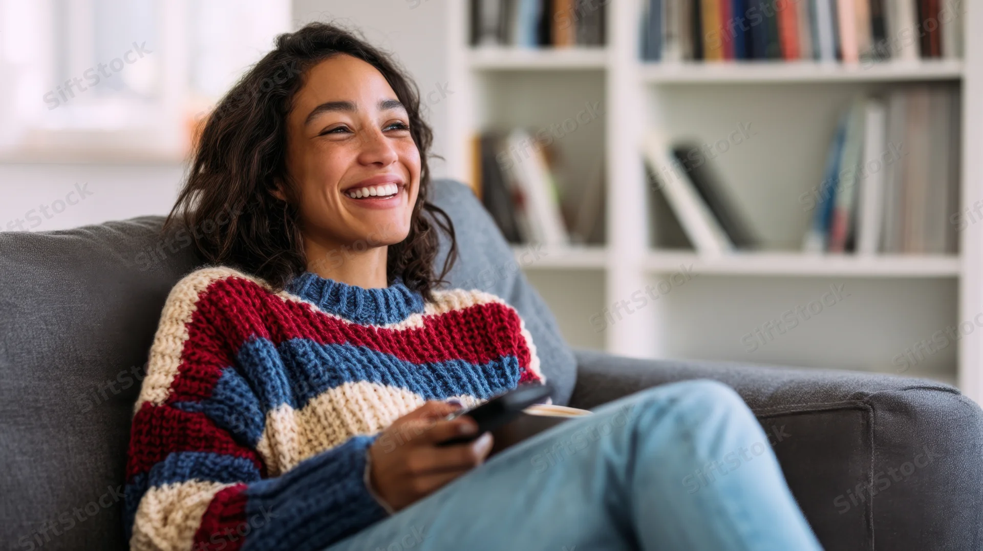 Young woman laughing while sitting crosslegged on a gray sofa holding a remote in a cozy living room with bookshelf in background