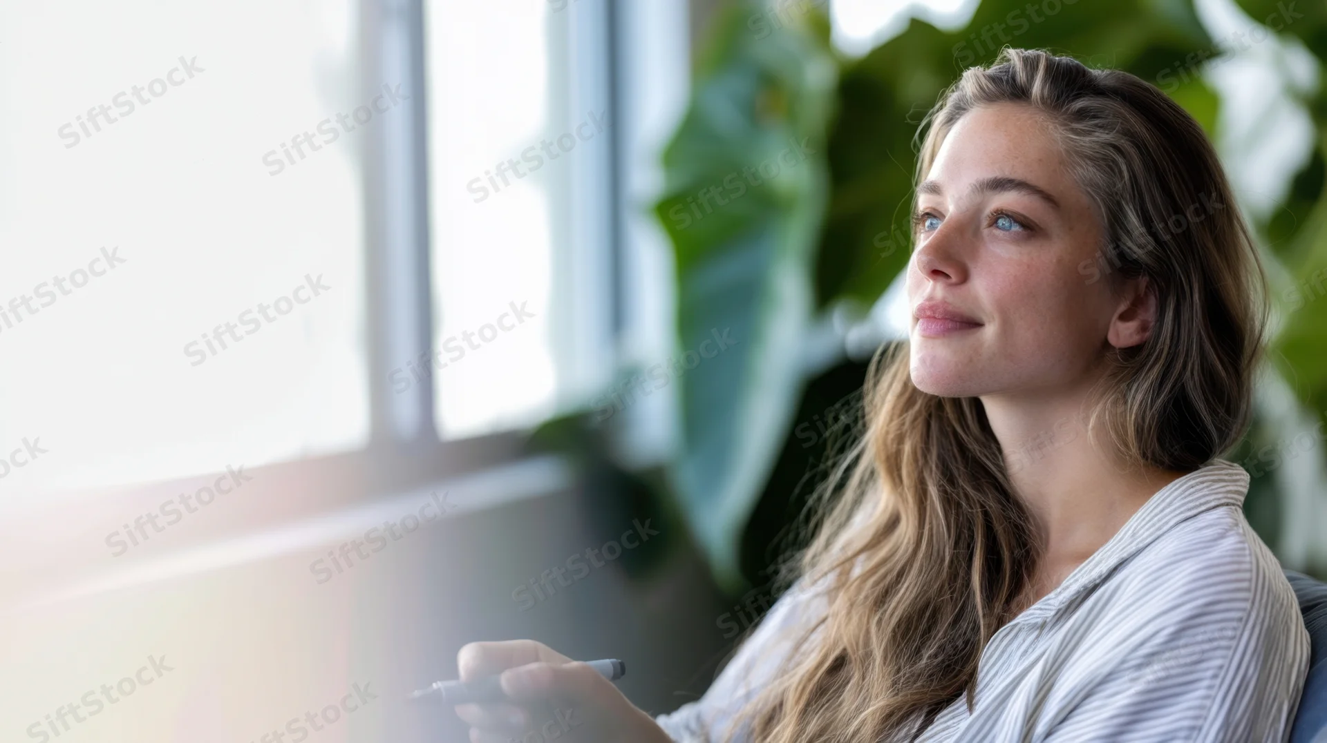 Young woman sitting in bright airy room holding pen and looking thoughtfully out window