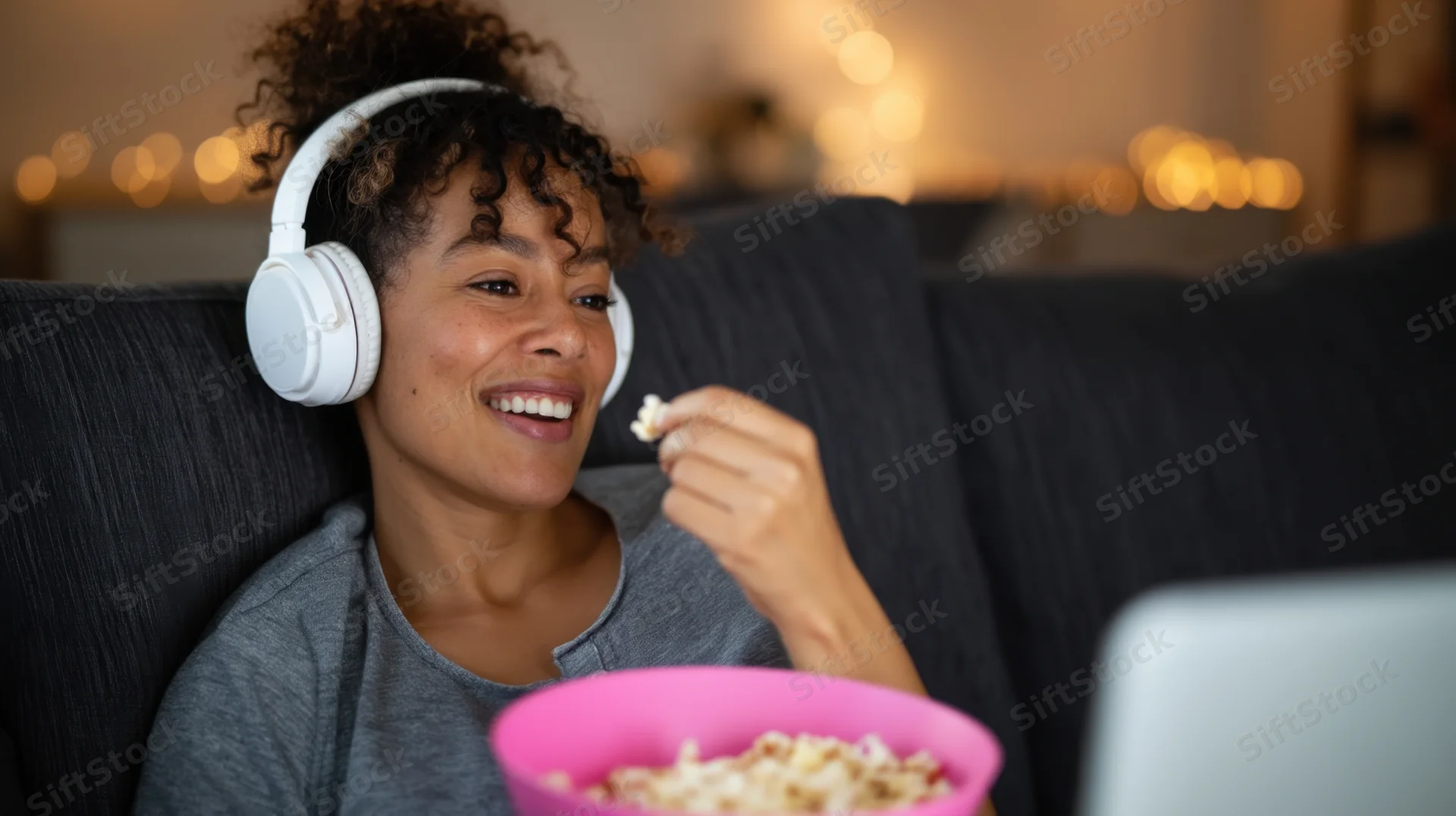 Woman smiling while eating popcorn and wearing headphones on a couch with a laptop nearby
