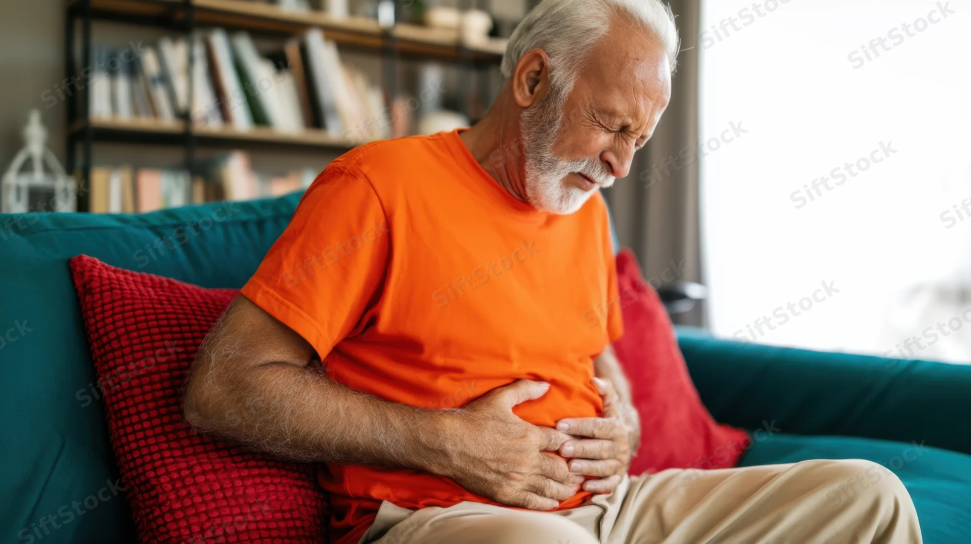 Elderly man clutching his stomach in pain while sitting on a teal couch at home