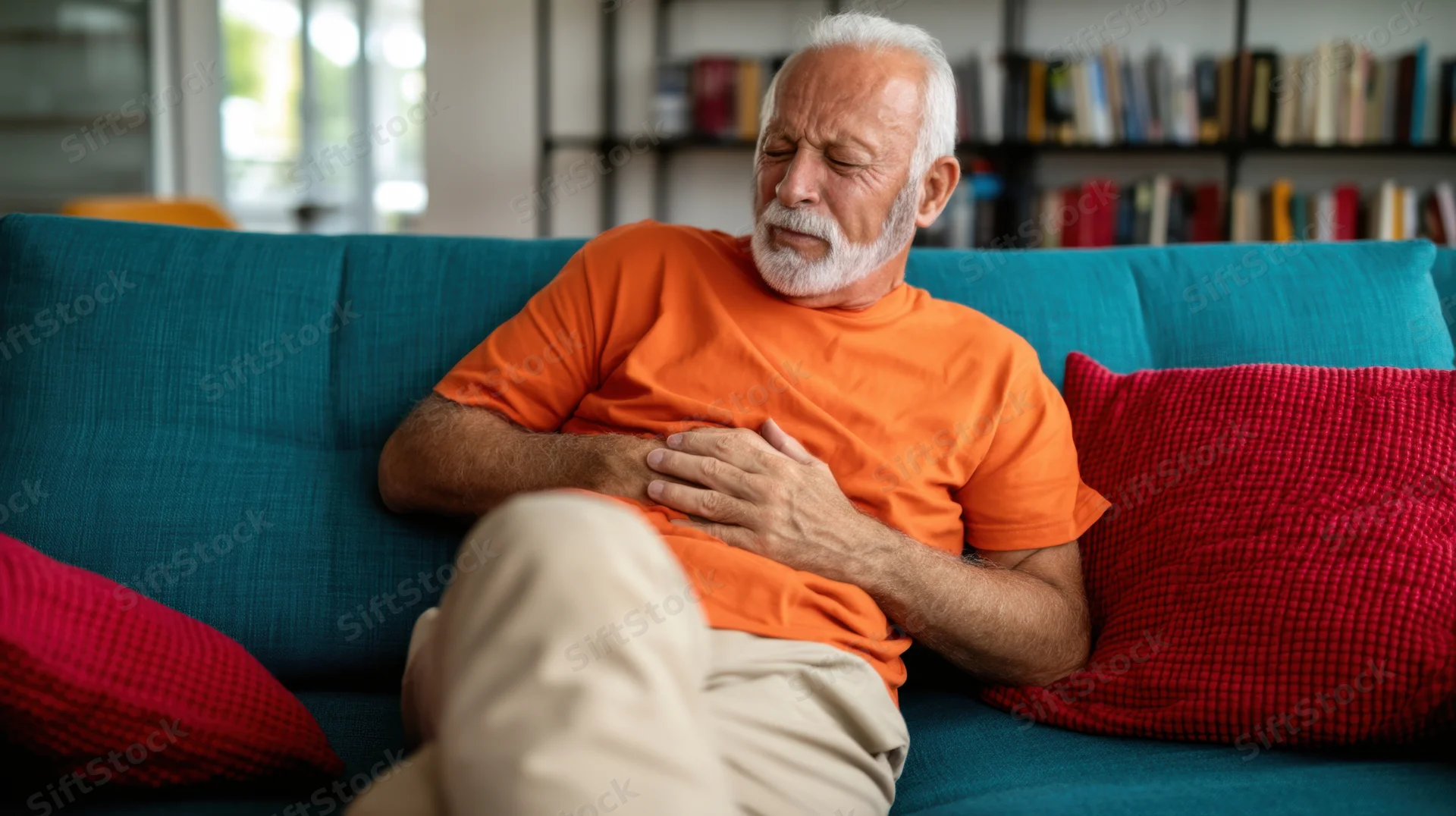 Elderly man holding his stomach in discomfort while sitting on a teal couch in a living room