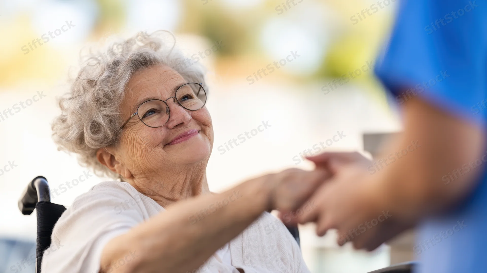 Elderly woman in wheelchair smiling while holding hands with caregiver outdoors