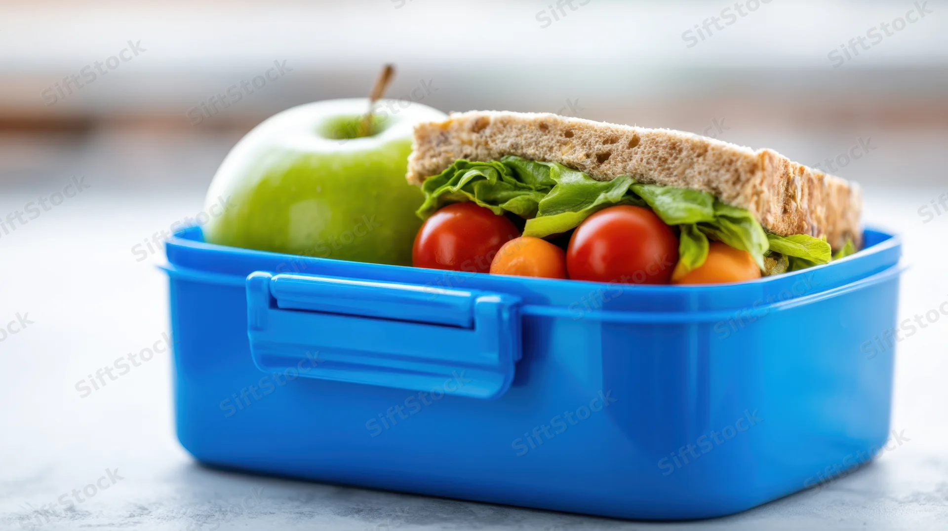 Blue lunchbox containing sandwich with lettuce and tomatoes, green apple, and small vegetables arranged neatly on a light surface