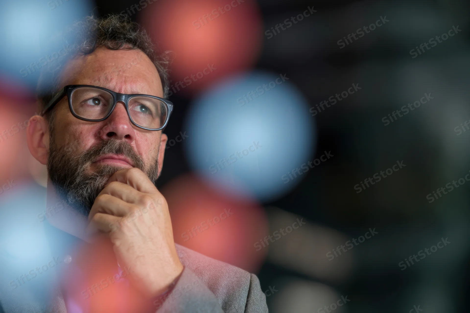 Businessman in suit with hand on chin contemplating in office setting with blurred lights in background