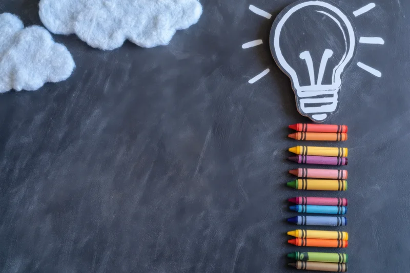 Colorful crayons arranged in ladder shape on chalkboard with lightbulb and clouds