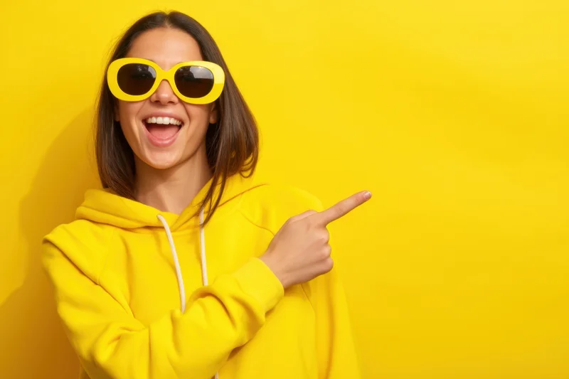 Young woman in yellow hoodie and oversized yellow sunglasses laughing and pointing to the side against yellow background