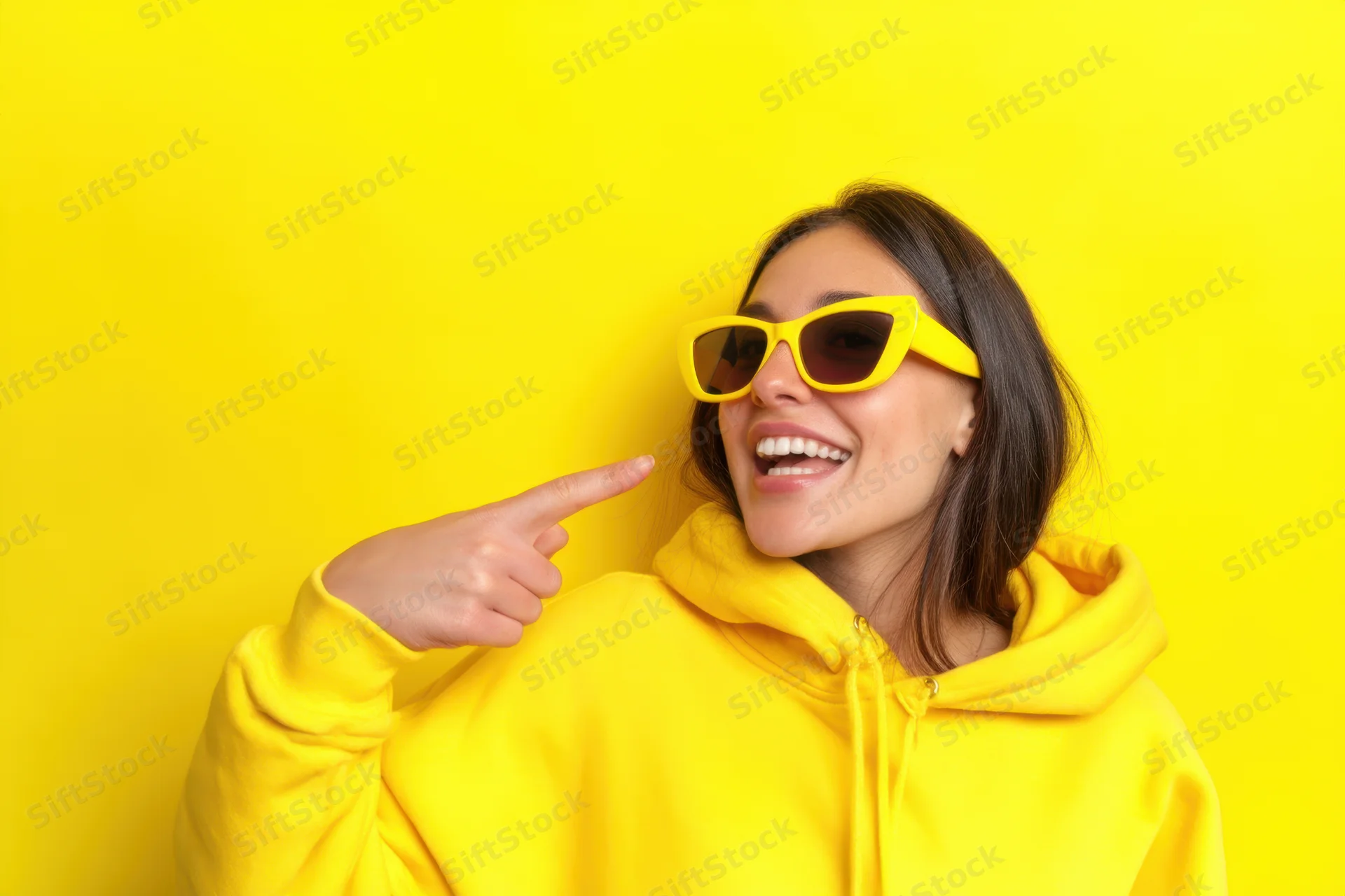 Young woman in yellow hoodie and sunglasses laughing while pointing to her face against yellow background
