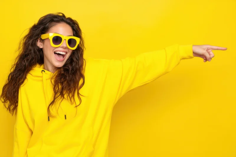 Young woman in yellow hoodie and sunglasses laughing and pointing against yellow background