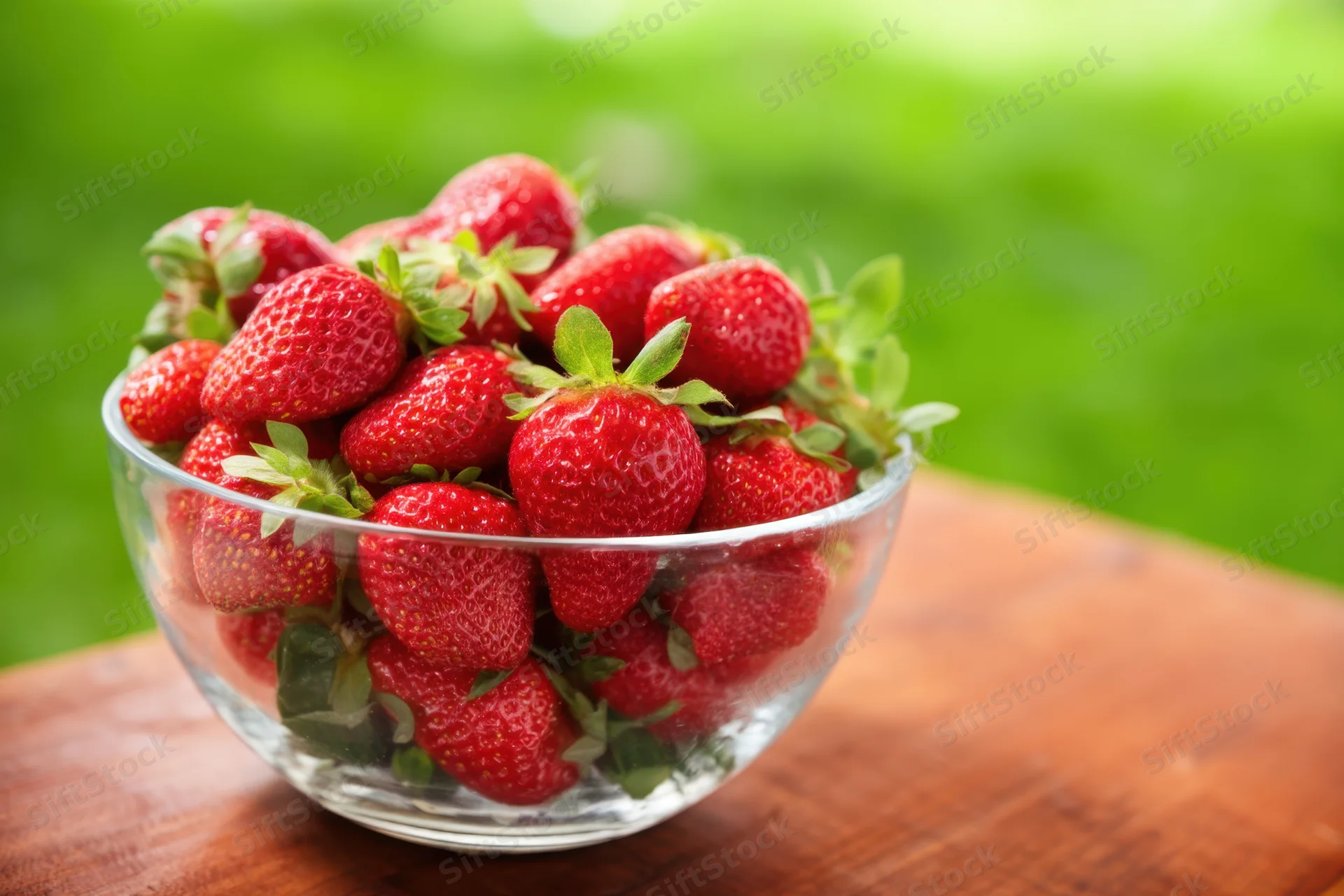 Fresh ripe strawberries in a clear glass bowl on a wooden table outdoors