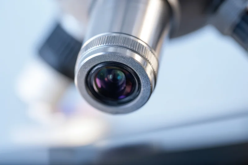Close-up of a microscope eyepiece lens with visible glass optics and metallic housing, set against a blurred laboratory background