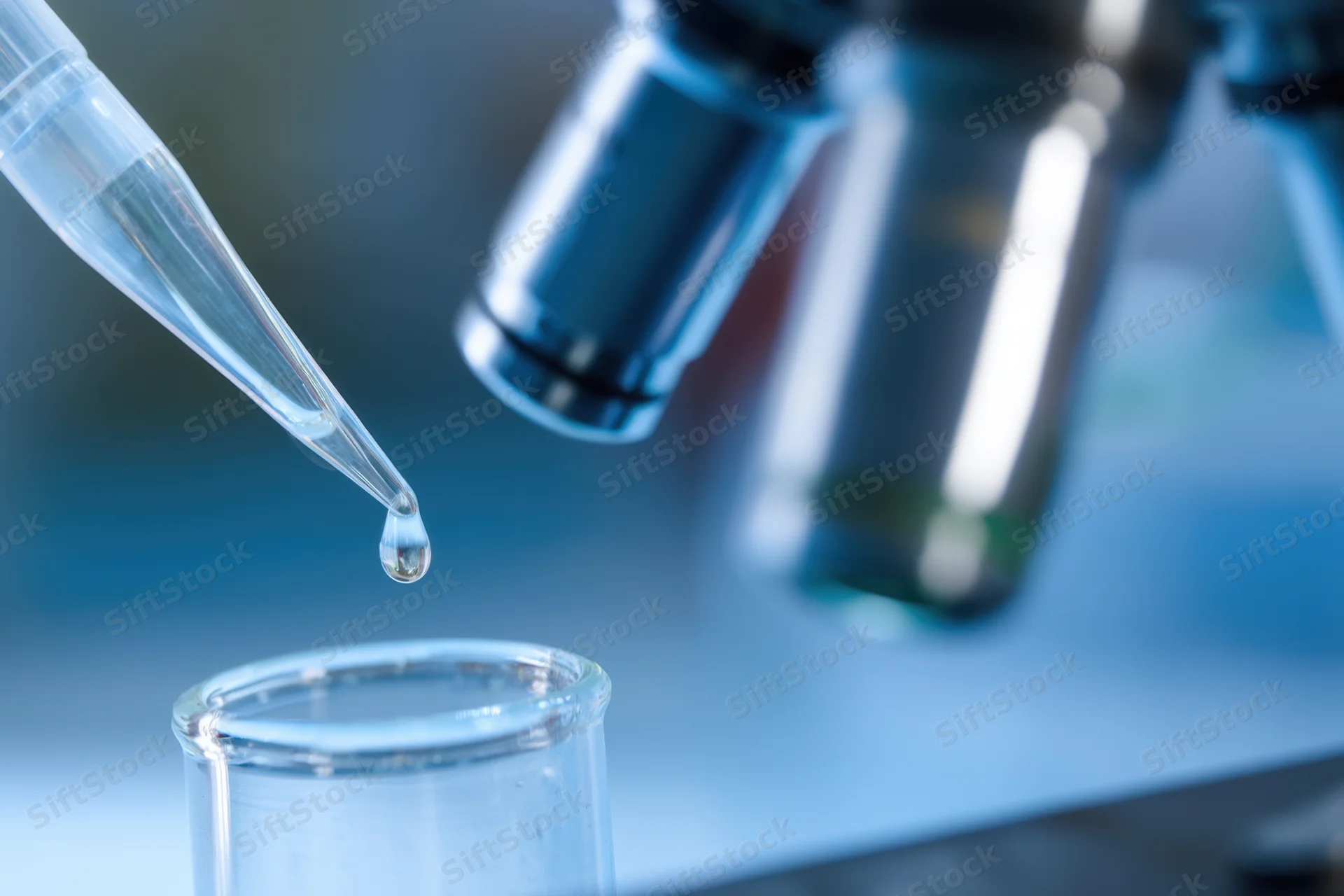 Pipette releasing a drop of liquid into a test tube in a laboratory setting with microscope in background