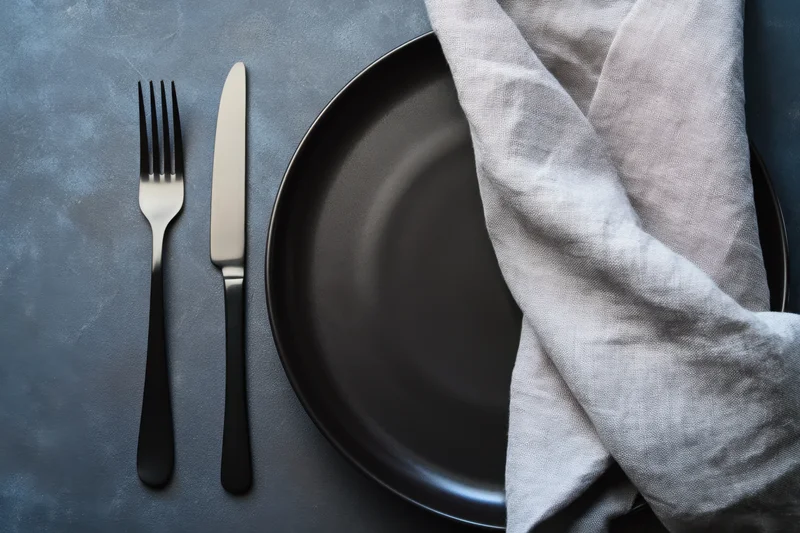 Black plate with fork, knife and napkin arranged on dark textured tabletop