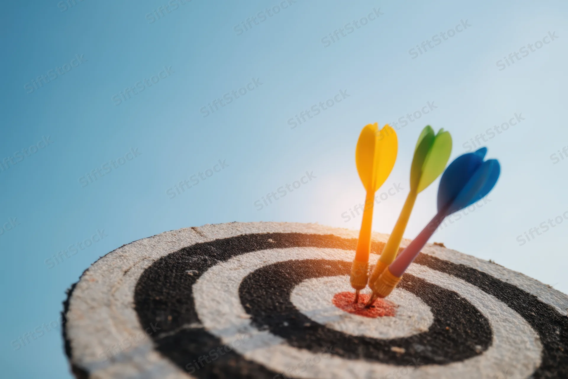 Three colorful darts hitting the bullseye of a wooden dartboard against a clear blue sky