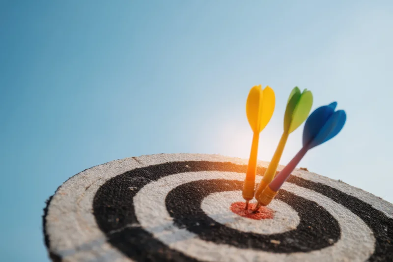 Three colorful darts hitting the bullseye of a wooden dartboard against a clear blue sky