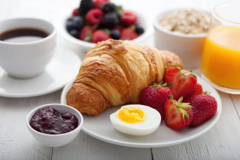 Breakfast spread with croissant, strawberries, boiled egg, coffee, orange juice, jam, and oatmeal on a white wooden table
