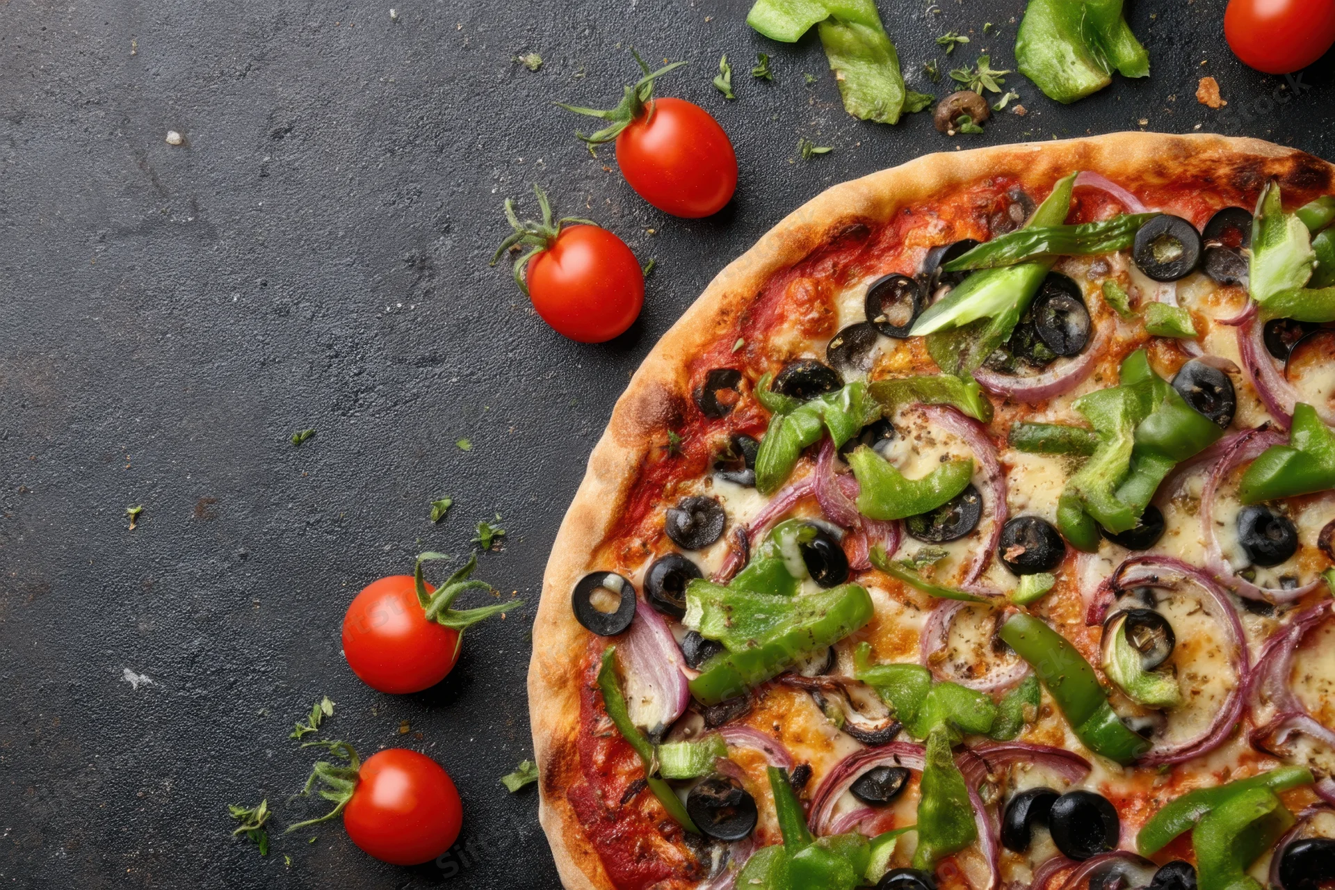 Half a vegetable-topped pizza on a dark surface with cherry tomatoes and peppers scattered around