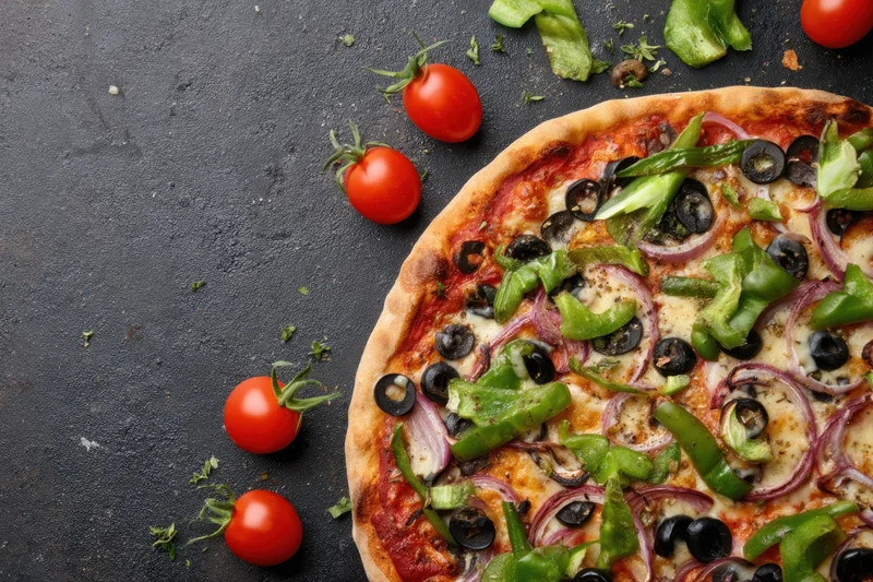 Half a vegetable-topped pizza on a dark surface with cherry tomatoes and peppers scattered around
