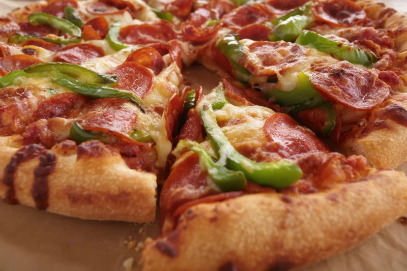Freshly baked pizza with golden crust topped with pepperoni and green peppers, sliced and arranged on a wooden surface