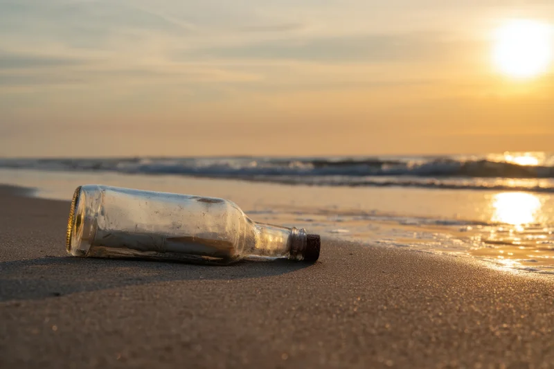 Glass bottle with cork lying on sandy beach at sunset near ocean waves
