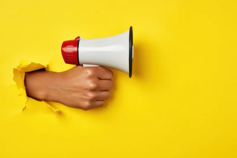 Hand breaking through yellow paper wall holding megaphone