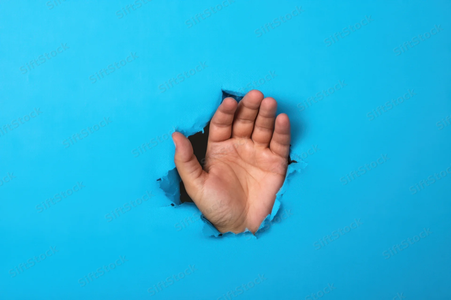 Hand reaching through a torn hole in a bright blue paper background