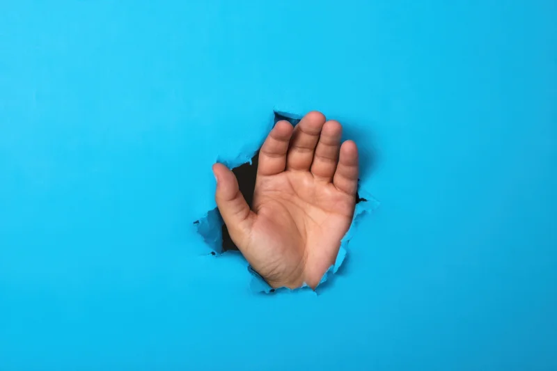 Hand reaching through a torn hole in a bright blue paper background