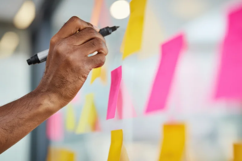 Hand writing with marker on transparent board surrounded by colorful sticky notes in office workspace