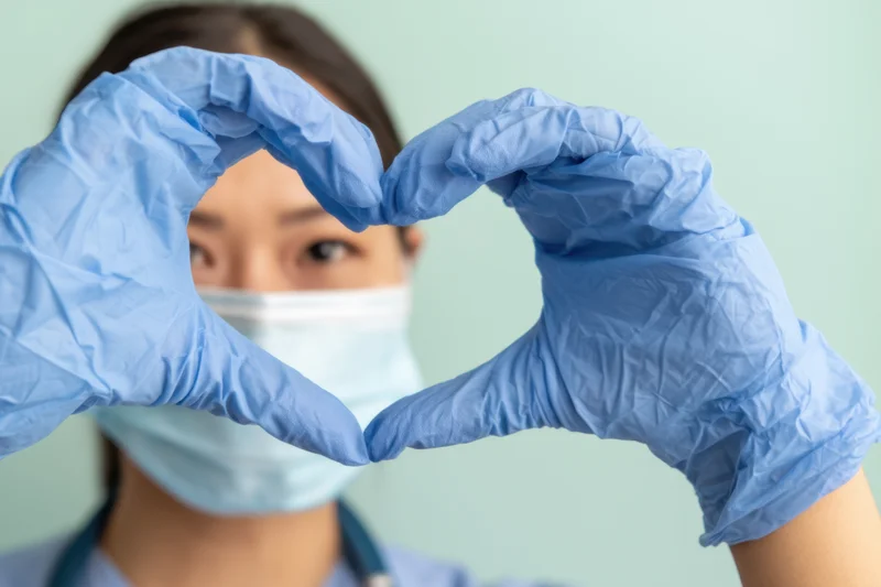 Woman in medical scrubs wearing face mask and blue gloves forming heart shape with hands