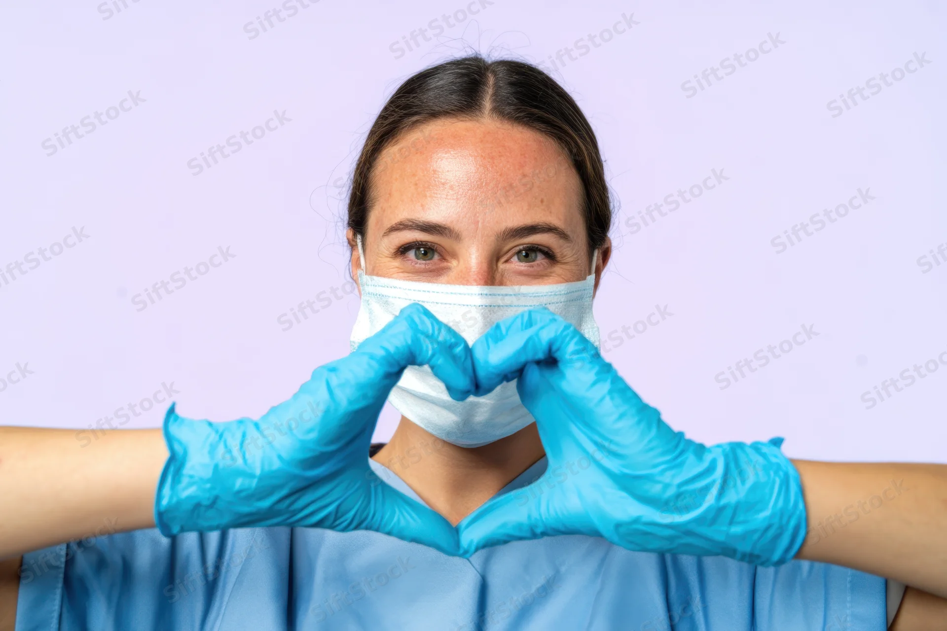 Woman in medical scrubs wearing face mask and blue gloves making heart shape with hands against light purple background