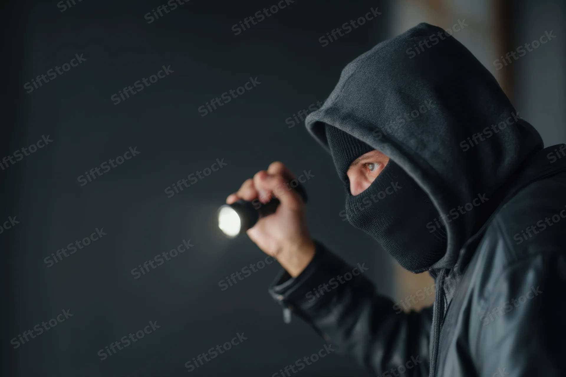 Hooded figure holding flashlight in dark environment, wearing black ski mask and hoodie, scanning surroundings cautiously