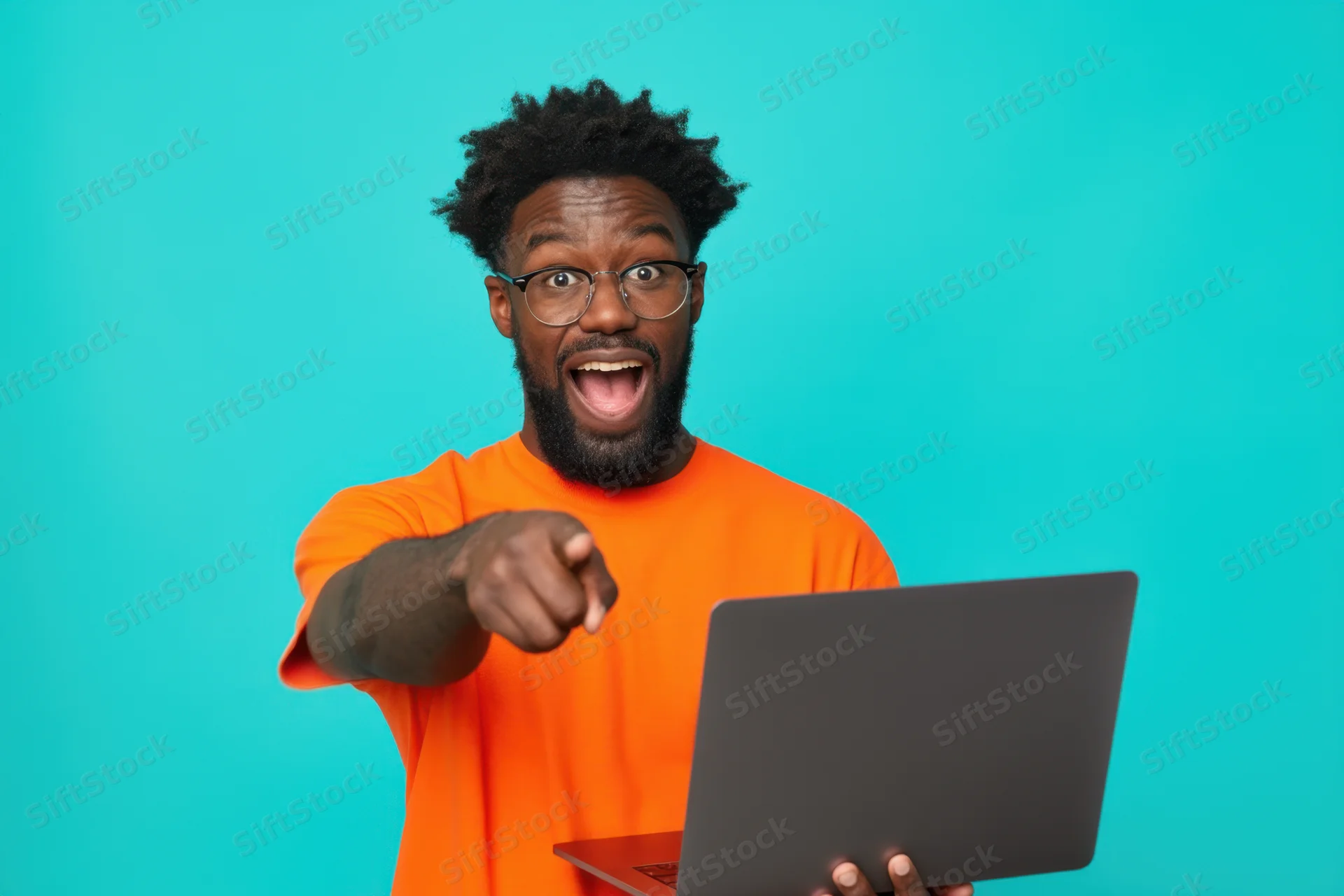 Man with beard and glasses in orange shirt pointing excitedly while holding laptop against teal background