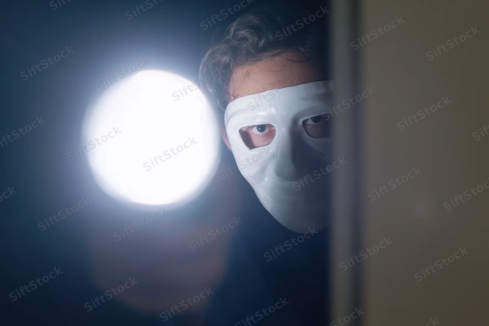 Masked figure peering through narrow opening with bright light in dark space