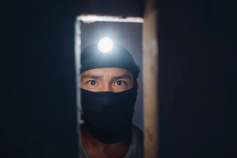 Man with headlamp and black face covering peering through narrow opening in dark environment