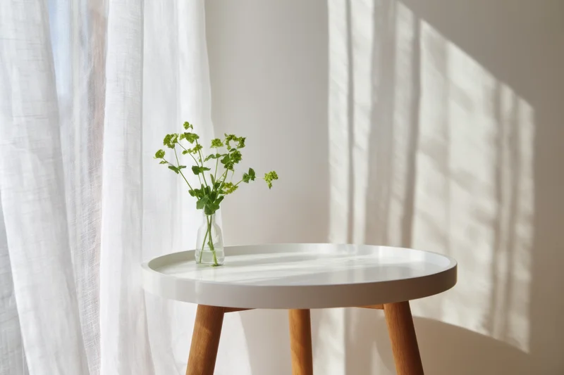 White round table with glass vase holding green plant stems, positioned near sheer curtain with sunlight casting shadows on wall