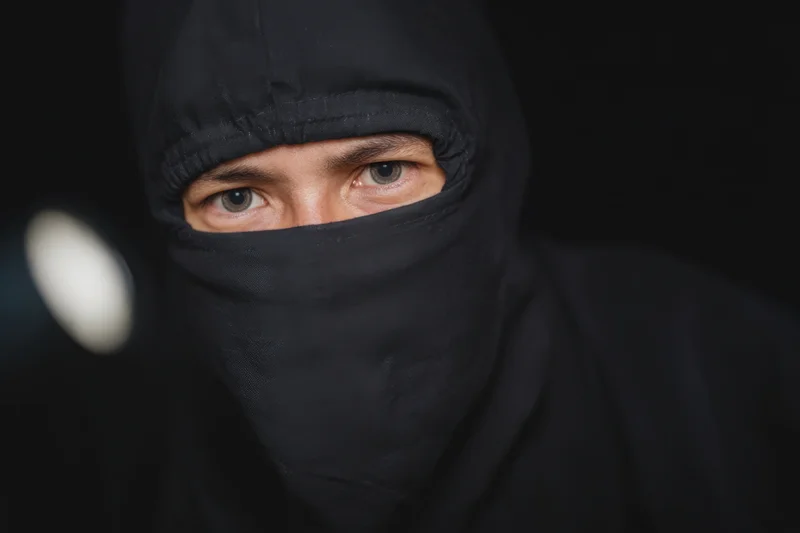 Man in black ninja outfit with hood and mask, eyes visible, staring intensely in dark setting