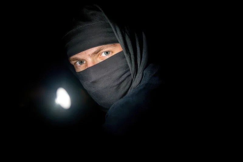 Ninja in black hooded outfit with flashlight beam in darkness, eyes visible through mask