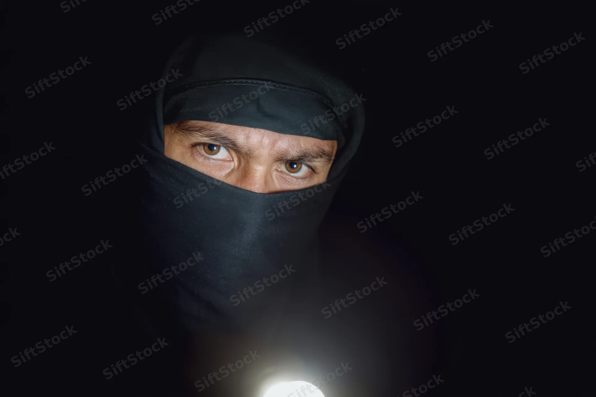 Hooded man in black ninja outfit holding flashlight in dark, eyes intensely focused on viewer