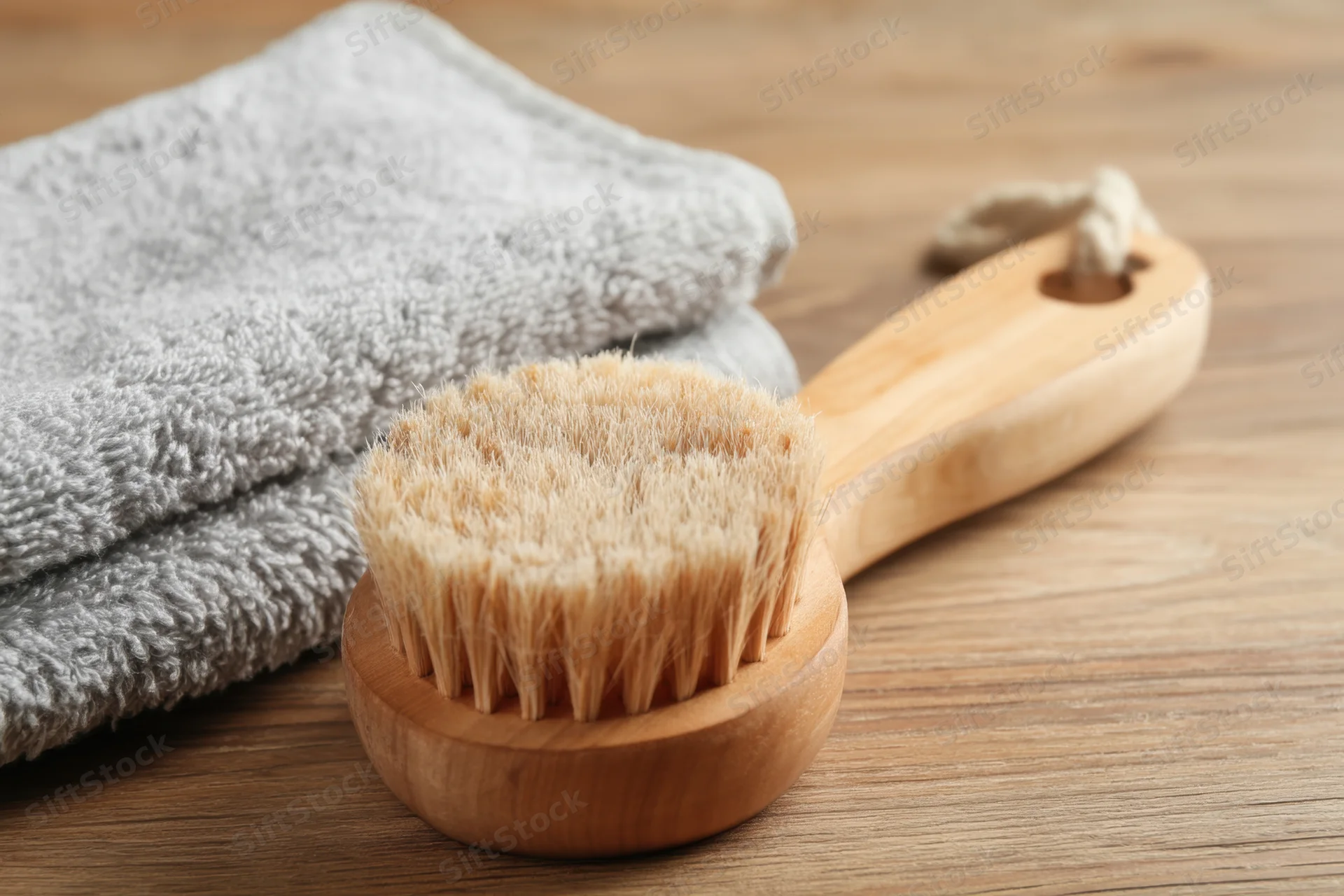 Wooden bath brush with soft bristles lying on a wooden surface beside a folded gray towel