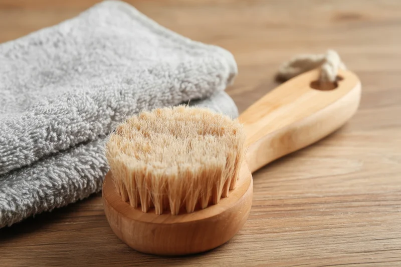Wooden bath brush with soft bristles lying on a wooden surface beside a folded gray towel