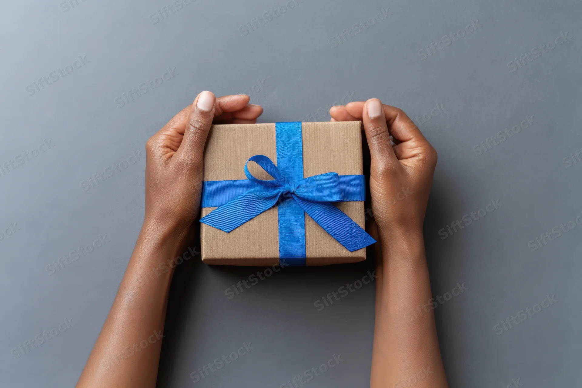 Hands holding a gift box wrapped in brown paper with a blue ribbon against a gray background