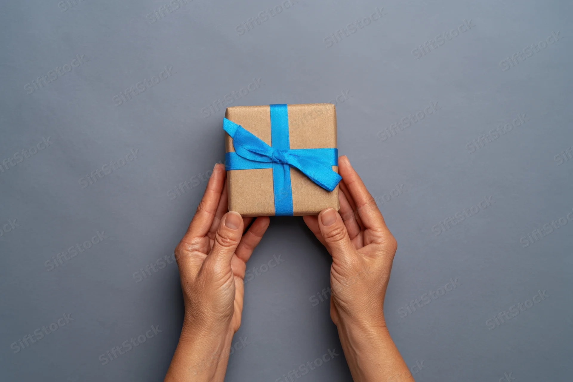 Hands holding a small gift box wrapped in brown paper with a blue ribbon against a gray background