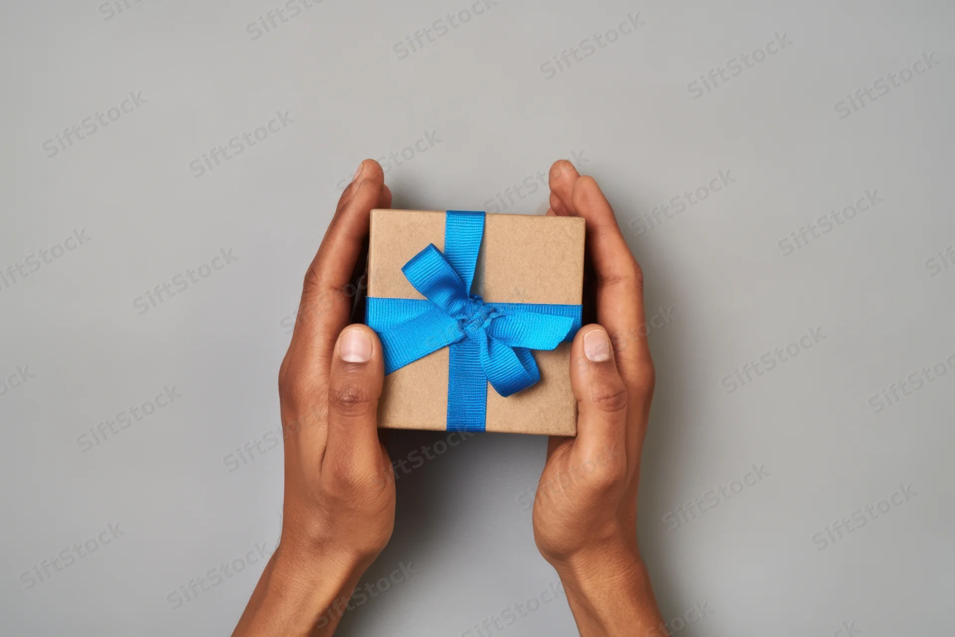 Hands holding a small gift box wrapped in brown paper with a blue ribbon against a gray background
