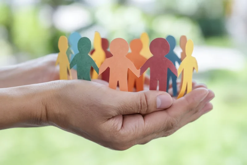 Hands cradling colorful paper cutouts of people in a circle outdoors with blurred green background