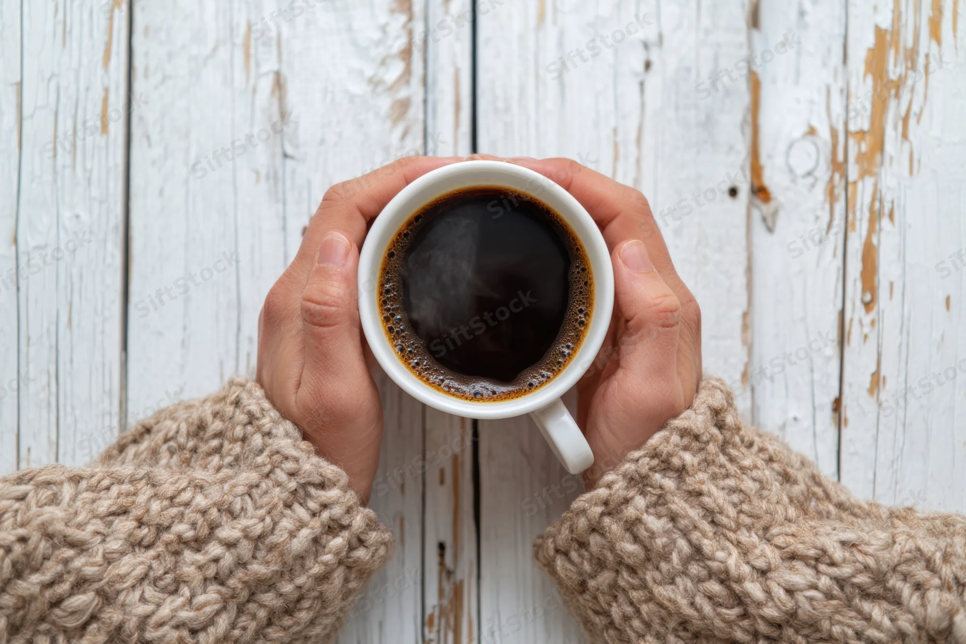 Hands holding a steaming cup of black coffee on a rustic white wooden table