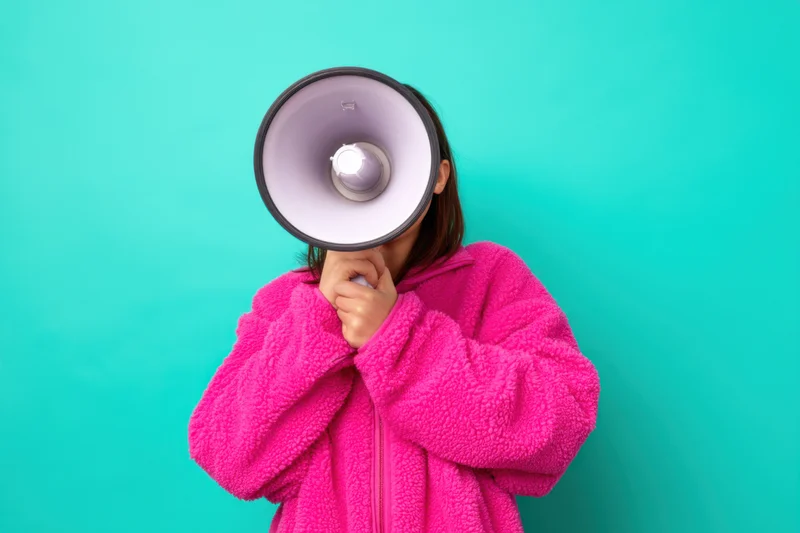 Woman in bright pink fluffy jacket holding megaphone to her mouth against teal background