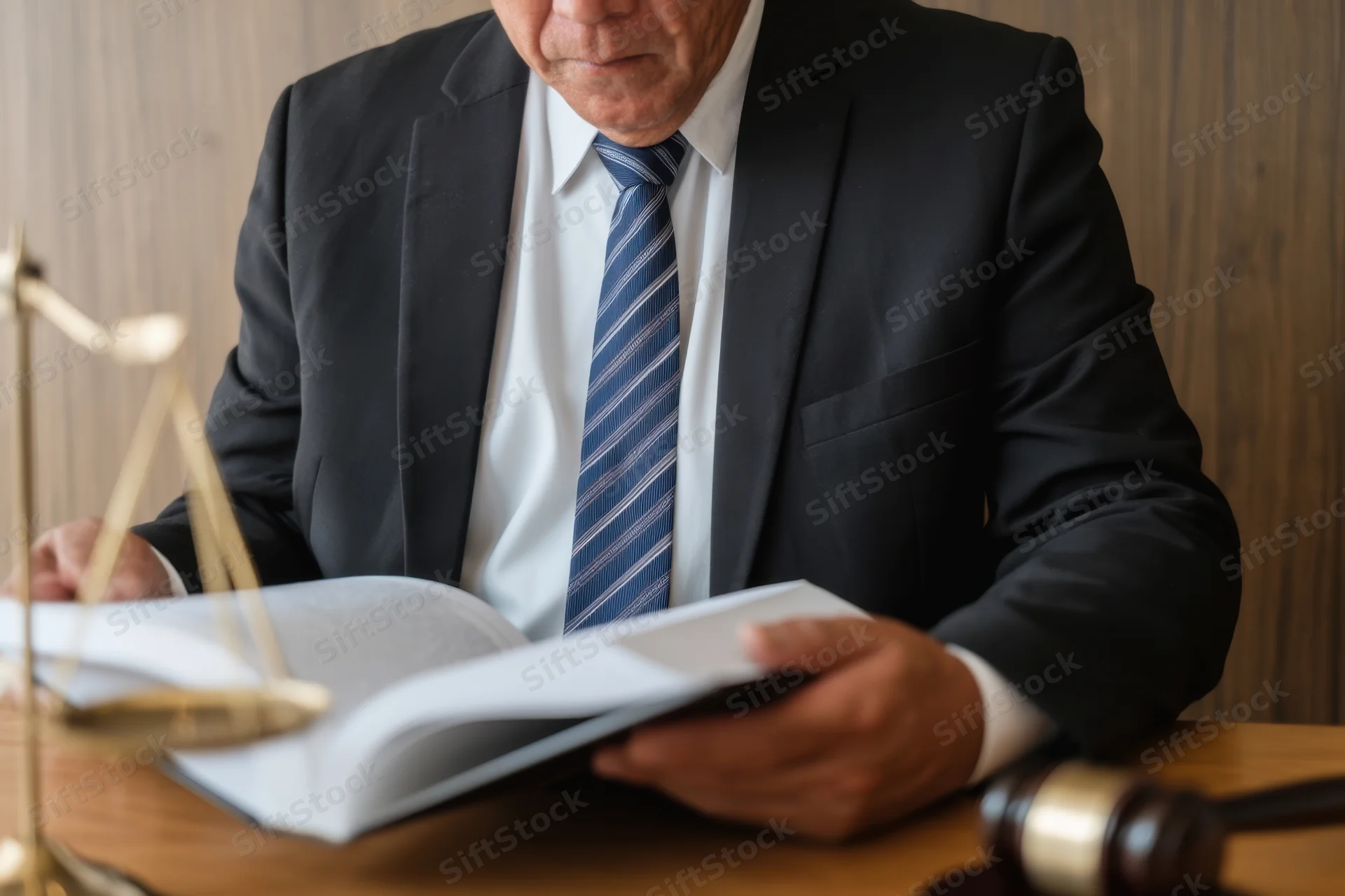 Lawyer in suit reading legal document at wooden desk with gavel and scales nearby