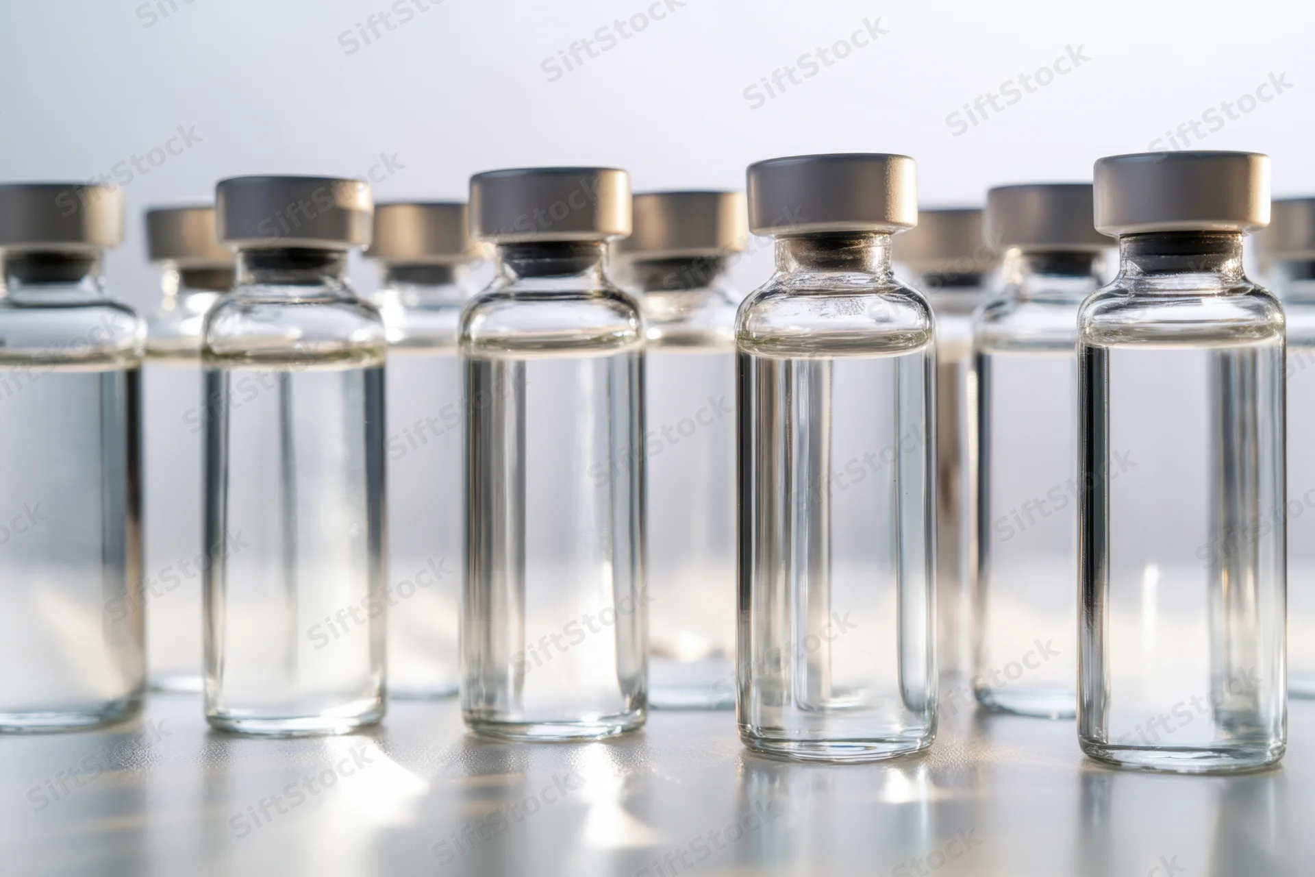 Row of glass vials filled with clear liquid capped and arranged on reflective surface