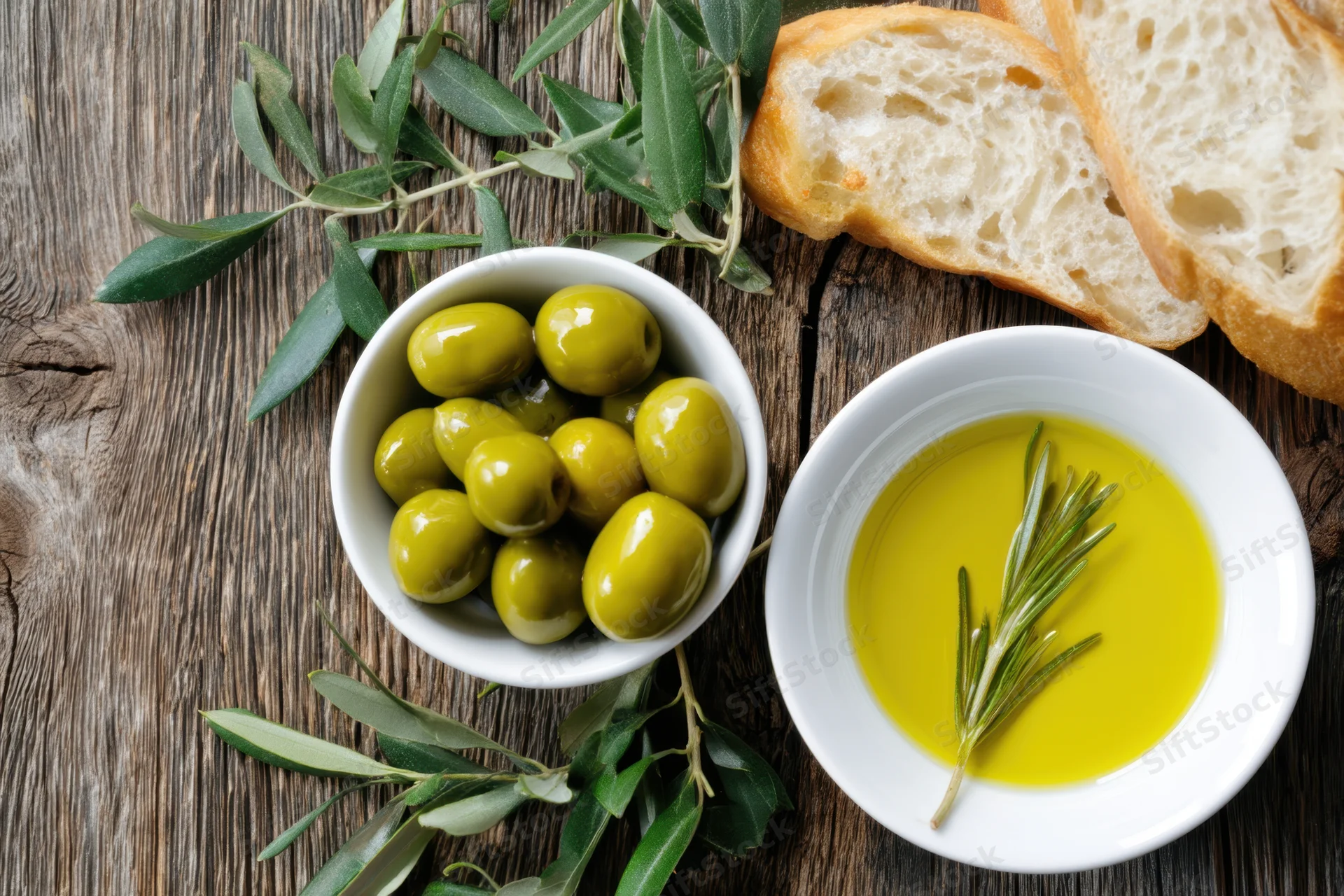 Green olives and olive oil with rosemary in white bowls beside bread on a rustic wooden table