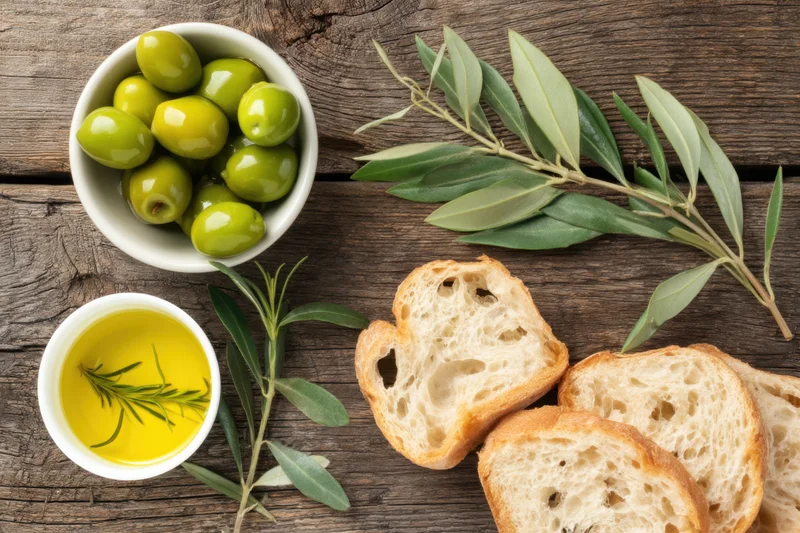 Slices of artisan bread, a bowl of green olives, and olive oil with rosemary sprigs arranged on a rustic wooden table