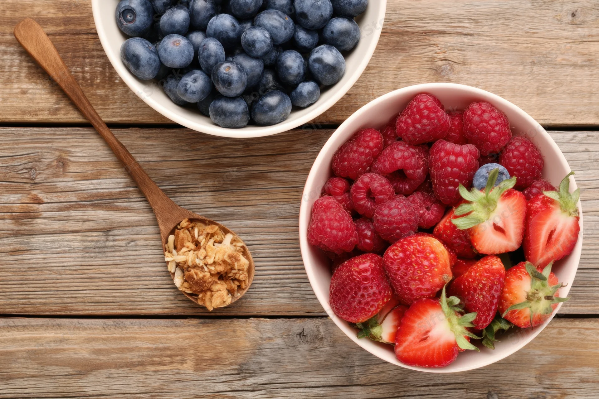 Two bowls of fresh berries and granola on a rustic wooden table with a wooden spoon beside them